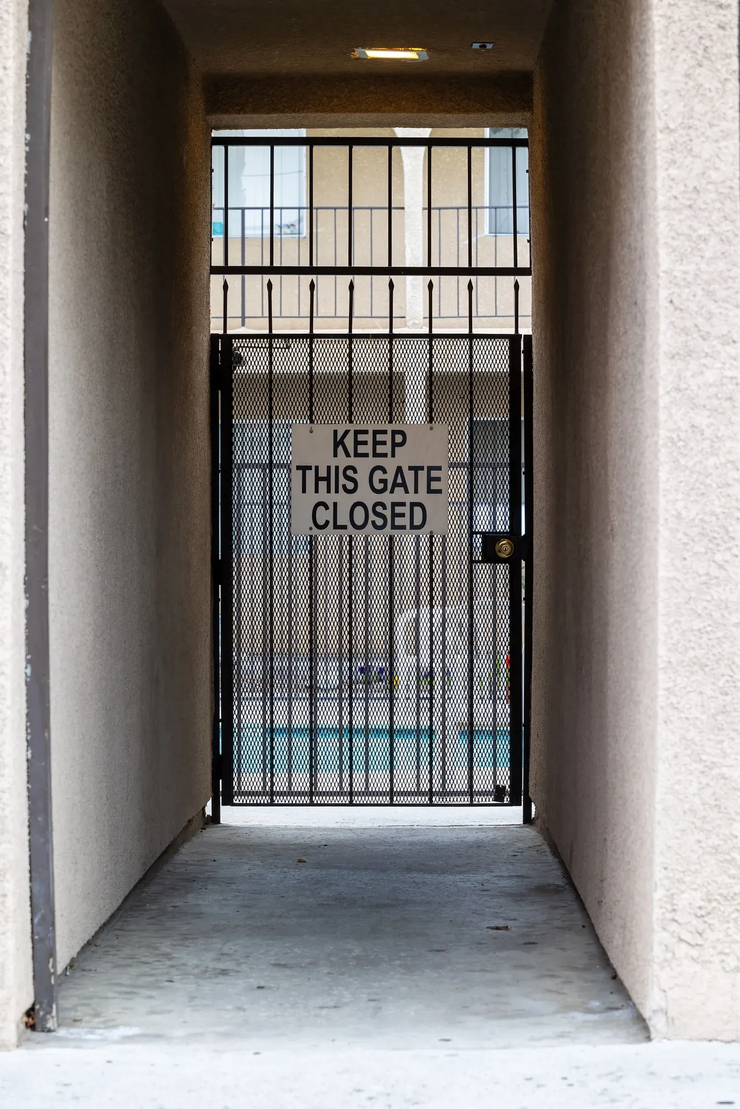 Black metal security gate at the end of a walkway between two walls, with a sign reading keep this gate closed and a pool visible beyond